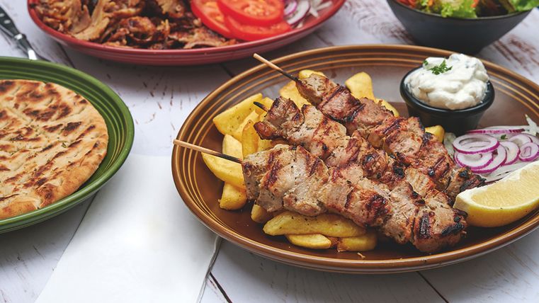 Sur une table en bois, il y a quatre plats : gyros avec frites, viande, pain et une salade mixte.