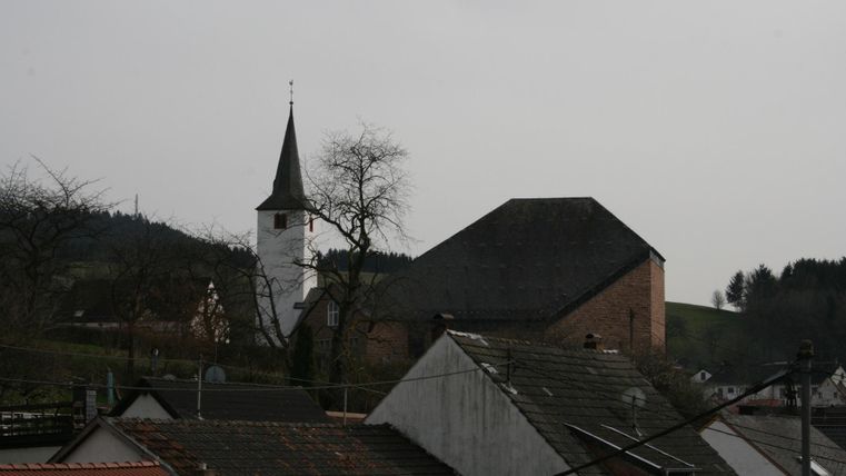 Eine kleine Kirche mit einem spitzen Turm steht neben einem Gebäudekomplex. Im Hintergrund sind Hügel und Bäume sichtbar.