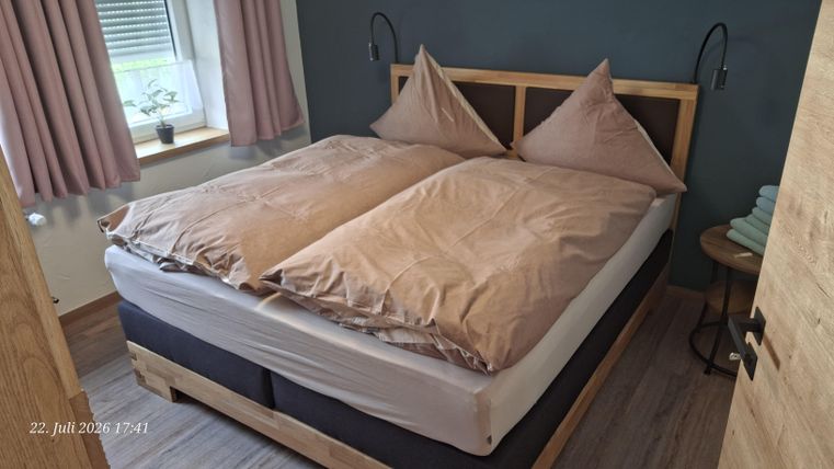 Ein gemütliches Schlafzimmer mit einem großen Doppelbett und beigen Bettdecken. Die Wände sind in einem dunklen Farbton gehalten, und es gibt Fenster mit hellen Vorhängen.