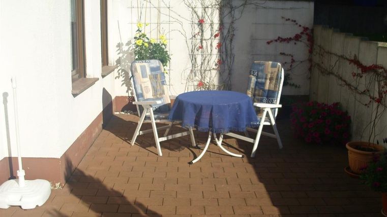 Ein gemütlicher Außenbereich mit einem runden Tisch und zwei Stühlen. Die gepflasterte Terrasse ist von Pflanzen umgeben und von Sonnenlicht durchflutet.
