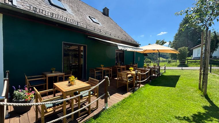Ein gemütlicher Außenbereich mit Holztischen und Stühlen, umgeben von grünem Rasen. Ein Sonnenschirm bietet Schatten an einem sonnigen Tag.