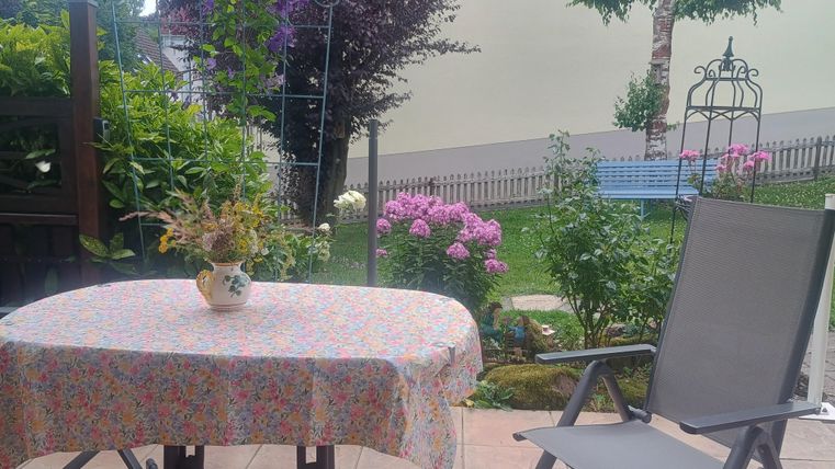 Ein gemütlicher Außenbereich mit einem Tisch und einem Stuhl. Umgeben von blühenden Pflanzen und einem gepflegten Garten.