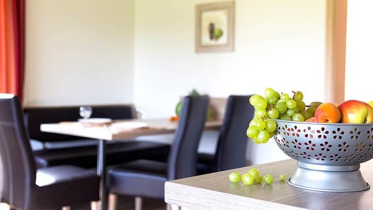 Eine gemütliche Küchenzeile mit einem Obstkorb voller Äpfel und Trauben im Vordergrund. Im Hintergrund ist ein Esstisch mit Stühlen und einer warmen Farbtönung zu sehen.