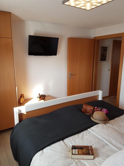 Ein modernes Schlafzimmer mit einem Doppelbett und hellem Holzschrank. An der Wand hängt ein Fernseher und ein Buch liegt auf der Bettdecke.