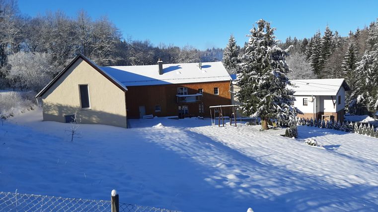 Ein verschneites Landschaftsbild mit einem modernen Haus und Bäumen. Der Himmel ist klar und blau, und der Schnee bedeckt die gesamte Umgebung.