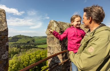 Ein Vater trägt seine Tochter auf dem Arm auf einer Trumspitze einer Burg. Das Mädchen zeigt freudig in die Ferne.