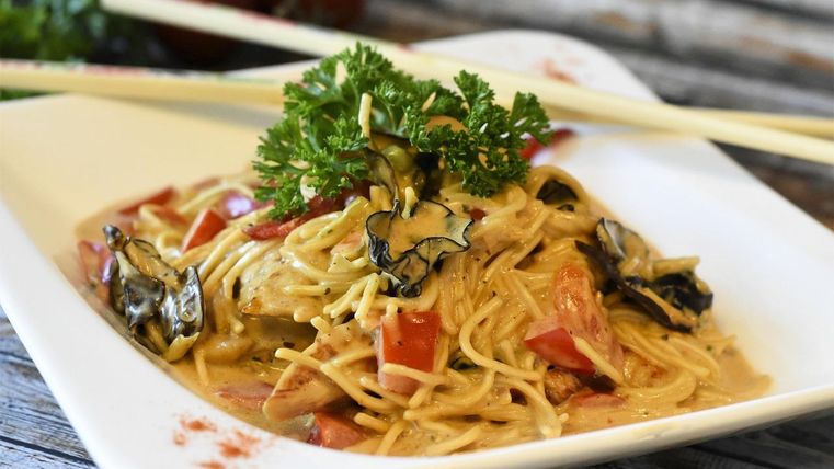 Ein Teller mit hohem Rand, auf dem ein asiatisches Nudelgericht mit verschiedenen Gemüsesorten und Kräutern angerichtet ist. Dazu liegen Essstäbchen auf dem Tellerrand.