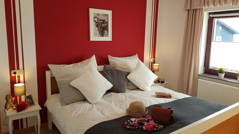 Ein gemütliches Schlafzimmer mit einem großen Bett und mehreren Kissen. Die Wände sind rot, und es gibt einen Fenster mit hellem Vorhang.