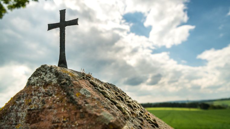 Ein kleines, schwarzes Kreuz auf einem moosbewachsenen Stein vor einem bewölkten Himmel.