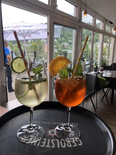 Zwei Cocktails mit Limetten- und Orangenscheiben auf einem Tablett in einem Bistro, im Hintergrund eine Terrasse mit Sonnenschirm.