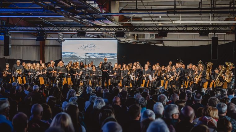 Ein prall gefüllter Raum aus Zuschauern vor einer großen Gruppe Musikanten und dem Dirigenten.