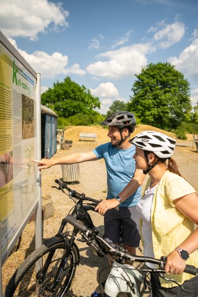 Zwei Personen auf Fahrrädern halten auf einem Rastplatz vor einer Informationstafel.