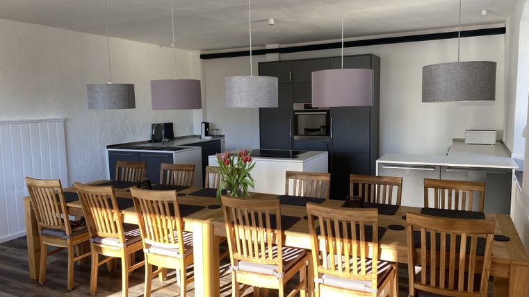 Ein modernes Esszimmer mit einem langen Holztisch und mehreren Stühlen. Die Küche im Hintergrund ist elegant und gut ausgestattet.