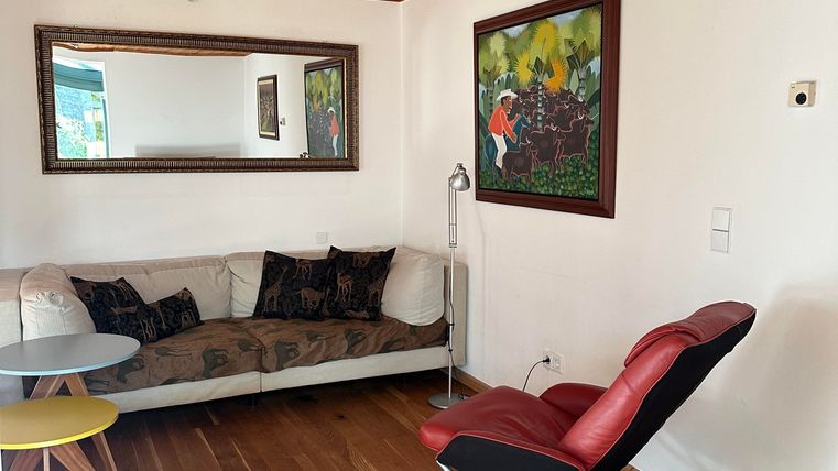 Ein helles Zimmer mit Holzboden und einem Sofa. An der Wand hängen Gemälde, und ein bequemer Stuhl steht im Raum.