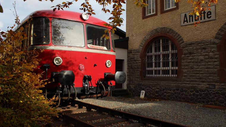 Ein roter Zug steht auf den Gleisen vor einem Bahnhofsgebäude. Die Umgebung ist von herbstlichen Blättern in warmen Farben umgeben.