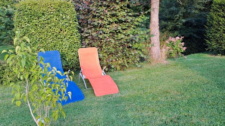 Zwei Liegen in einem grünen Garten, eine blau und eine orange. Umgeben von Bäumen und Sträuchern, ideal zum Entspannen.