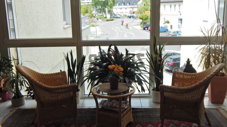 Gemütlicher Raum mit zwei Stühlen und einem Tisch, umgeben von Pflanzen. Durch das große Fenster blickt man auf eine ruhige Straße.