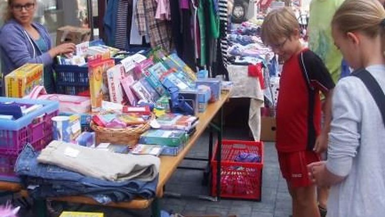 Ein Flohmarkt mit mehreren Ständen, die Bücher, Kleidung und Spielsachen verkaufen. Kinder und Erwachsene stöbern zwischen den Waren.