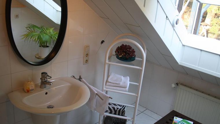 Ein modernes Badezimmer mit einem Waschbecken und einem großen Spiegel. Im Hintergrund sieht man einen Handtuchhalter und eine Pflanze, sowie Tageslicht von einem Fenster.