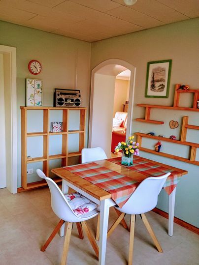 Ein gemütlicher Essbereich mit einem Holztisch, weißen Stühlen und einer bunten Tischdecke. Im Hintergrund sieht man Regale und einen alten Radio.