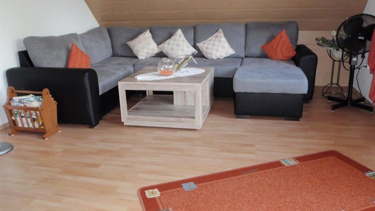 Ein gemütliches Wohnzimmer mit einer grauen Couch und orangefarbenen Kissen. Ein Couchtisch steht in der Mitte und ein Teppich liegt auf dem Boden.