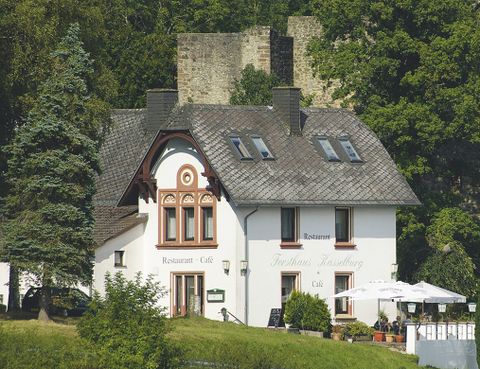 <p>Im Vordergrund sihet man ein weißes Haus mit der Aufschrift "Restaurant Café" und "Forsthaus Kasselburg", Davor stehen zwei große Schirme und Tische mit Stühlen, Im Hintergrund ist eine Burg und Bäume</p>