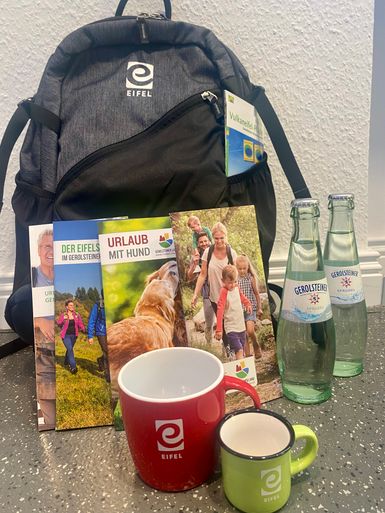 Ein Eifel-Rucksack mit mehreren touristischen Prospekten, zwei Eifel-Tassen und zwei Glasflaschen Gerolsteiner Sprudel.