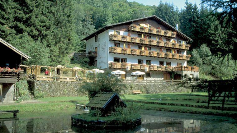 Un hôtel confortable au milieu des arbres verts et de la nature. Devant le bâtiment se trouve un petit étang avec un banc en bois et des parasols.
