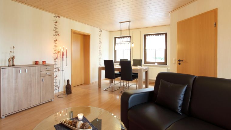 Ein modernes Wohnzimmer mit einem schwarzen Sofa und einem Glastisch. Im Hintergrund befinden sich ein Esstisch und ein Sideboard an der Wand.