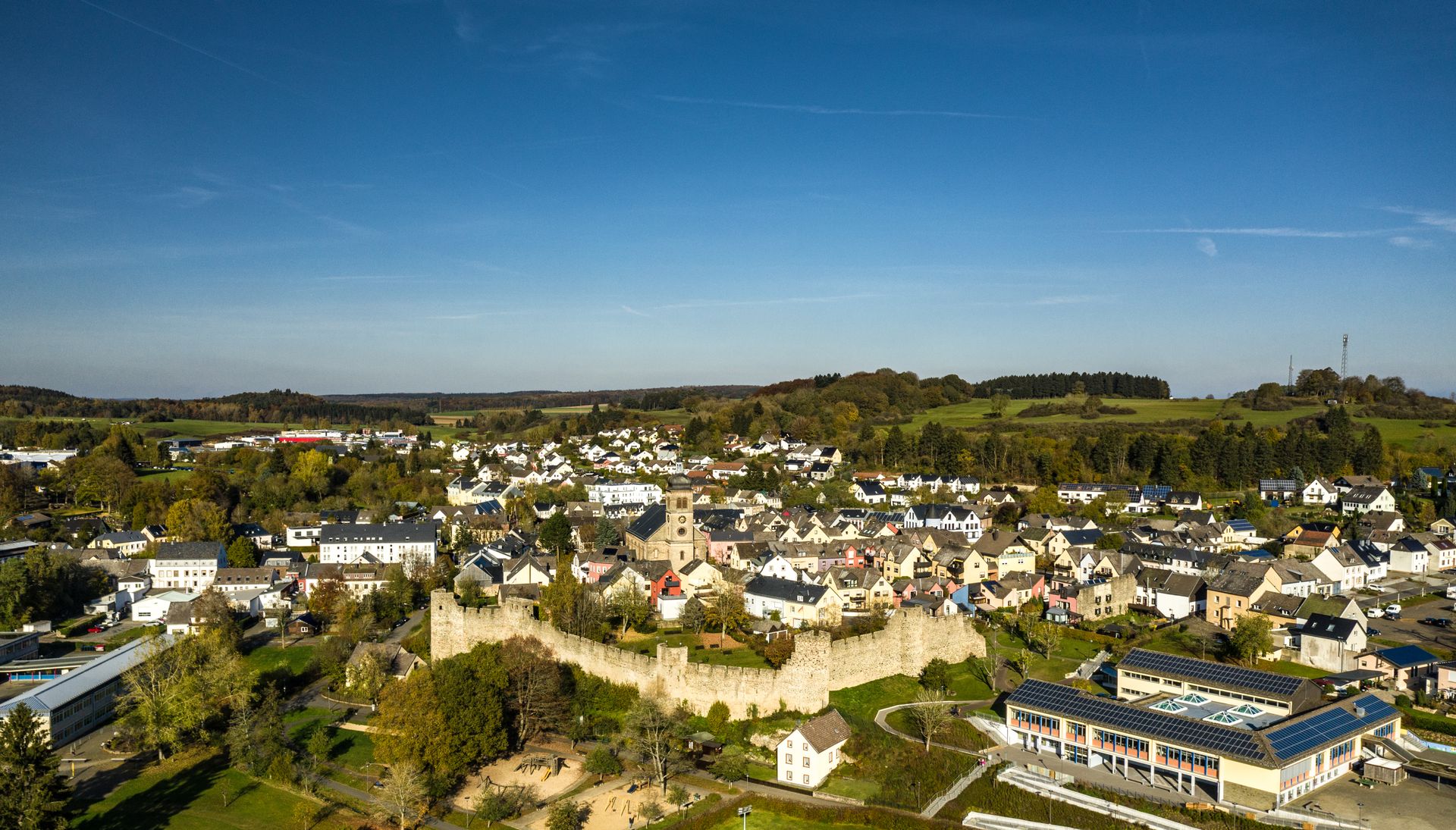 Luftaufnahme von Hillesheim mit historischer Stadtmauer und Kirche, umgeben von grüner Landschaft.