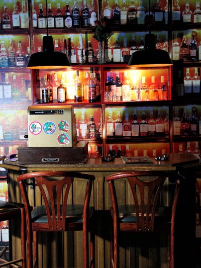 Un bar confortable avec des chaises en bois et une étagère pleine de spiritueux. La lumière crée une atmosphère accueillante.
