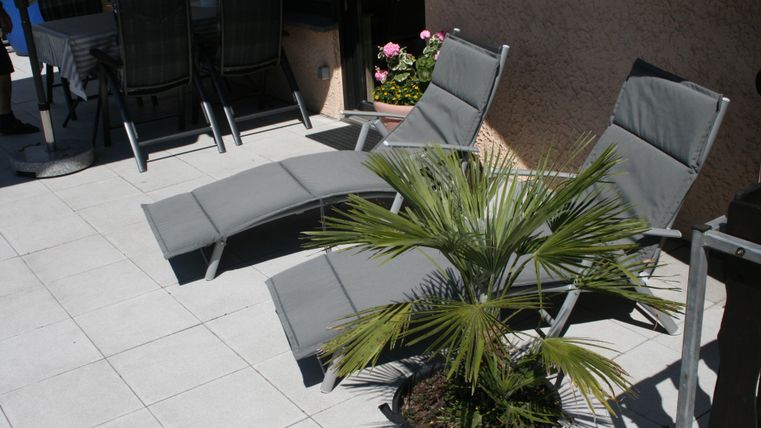 Ein gemütlicher Außenbereich mit zwei Liegen und einer Palme. Im Hintergrund sind ein Tisch und Stühle sichtbar.