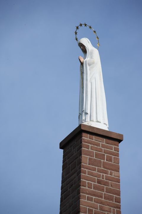 Eine Statue der Jungfrau Maria steht auf einem Ziegelturm. Der Himmel ist klar und blau.