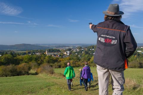 Eine Hut tragende Person steht mit dem Rücken zum Betrachter und zeigt mit einer Pistole in die Ferne. Sie trägt eine Jacke mit der Aufschrift "...durch Wald und Flur auf Eifel-Krimi-Spur" auf dem Rücken. Vor der Person liegt eine große Wiese und dahinter in der Ferne ein Dorf mit vielen Häusern.