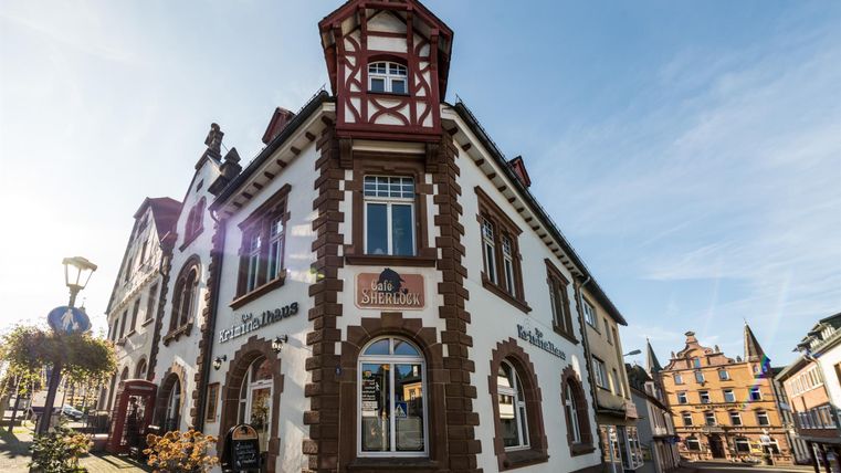 Das Café Sherlock und Kriminalhaus in Hillesheim zeigt eine historische Fassade mit Fachwerkelementen. Ein Straßenschild und Blumen schmücken den Gehweg.