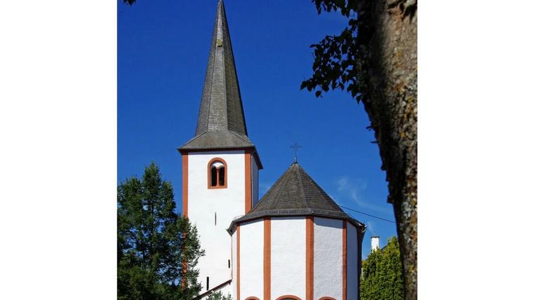 Eine alte Kirche mit einem hohen, spitzen Turm und einer runden, weiß gestrichenen Fassade. Umgeben von Bäumen und blauem Himmel.