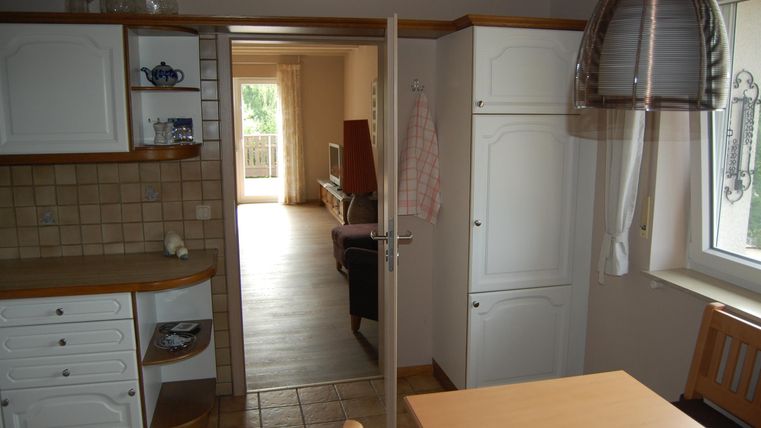 A bright kitchen with white cabinets and a table. In the background, a living area is visible.