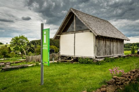 Grünes Infopunktschild vor einem Holzhäuschen auf einer grünen Wiese, bewölkter Himmel im Hintergrund.