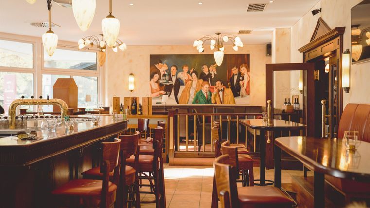 An elegant bar with wooden chairs and a large painting on the wall. The room is brightly lit and conveys an inviting atmosphere.