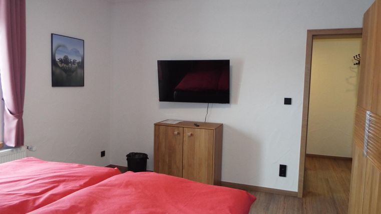 Ein gemütliches Schlafzimmer mit zwei roten Betten und einem Fernseher an der Wand. Die Wände sind hell, und es gibt ein Bild sowie einen Schrank im Raum.