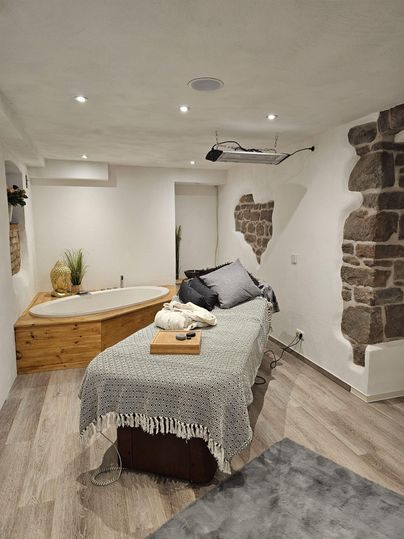 Une salle de bien-être confortable avec un lit douillet et une baignoire en bois. Des murs clairs et un éclairage doux créent une atmosphère relaxante.