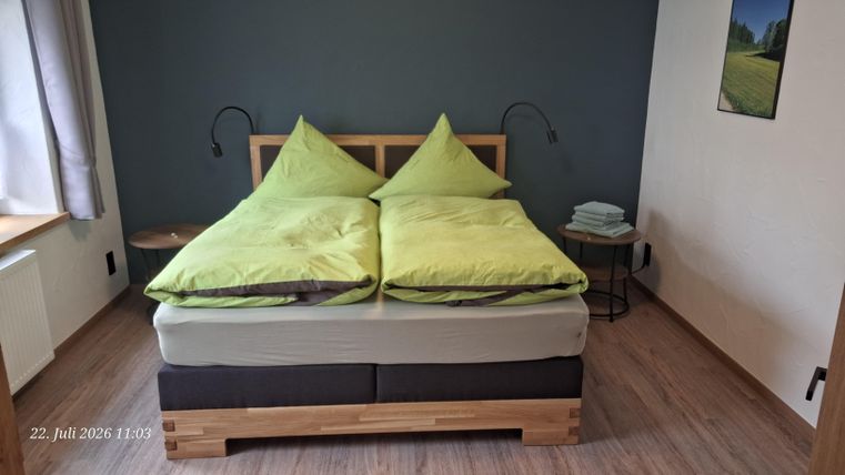 Ein gemütliches Schlafzimmer mit einem großen, bequemen Bett und grünen Kissen. Neben dem Bett befindet sich ein kleiner Nachttisch mit gestapelten Handtüchern.