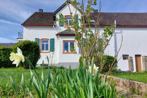 Een mooi huis met witte muren en kleurrijke raamkaders. Op de voorgrond groeien gele bloemen en groen gras.