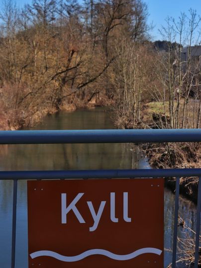 Een rustige rivier met een bord "Kyll" op de voorgrond. Op de achtergrond zijn bomen en een heldere blauwe lucht te zien.