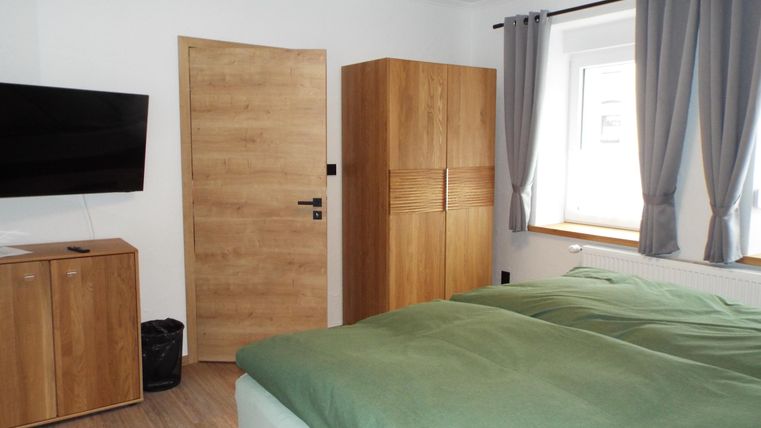 Ein modernes Schlafzimmer mit einem großen Bett und einer Holzkommode. Ein Fernseher hängt an der Wand und das Fenster lässt viel Tageslicht herein.