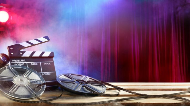 Filmrolle, Filmklappe und Kamera auf einem Tisch vor einem bunten, nebligen Hintergrund mit roten Vorhängen und Scheinwerfern.