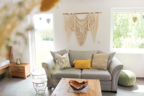 Ein helles Wohnzimmer mit einem grauen Sofa und dekorativen Kissen. An der Wand hängt eine kunstvolle Wanddekoration aus Makramee.