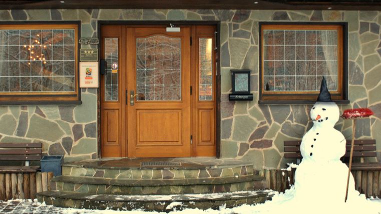 Une maison revêtue de bois avec une façade en pierre et de grandes fenêtres. Dehors, un bonhomme de neige se tient avec un chapeau noir et une pelle rouge.