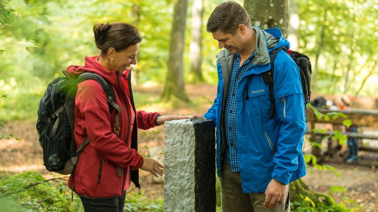 Zwei Personen stehen im Wald neben einem schwarz-weißen Grenzstein.