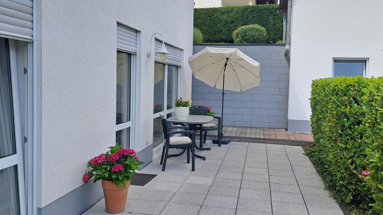 A cozy terrace with a table, chairs, and a parasol. Colorful flowers in a pot complement the outdoor area.